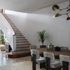 The dining area and staircase are viewed as a cohesive whole, showcasing the flow between social spaces. The material palette of wood, marble, and soft textiles, along with curated art and lighting, creates an atmosphere of understated elegance.