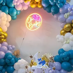 A dreamy "Oh Baby" baby shower setup. The focal point is a beautiful cake with a teddy bear on the moon, set against a circular balloon arch in shades of blue, purple, and gold, with a custom neon sign.