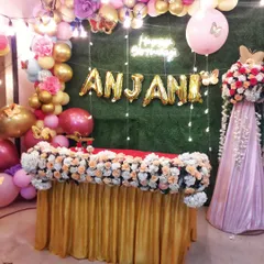 A birthday party cake-cutting table decoration. The setup includes a green wall backdrop, a 'Happy Birthday' neon sign, personalized name foil balloons, and a colorful balloon garland.
