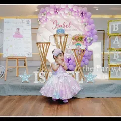 The birthday girl, Isabel, looks precious in front of her baby elephant-themed first birthday decor. The setup uses a soft lavender and white color palette with cute elephant illustrations and "BABY" blocks.