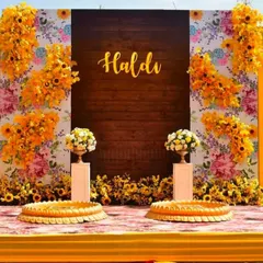 A close-up of a beautiful Haldi backdrop, combining a rustic wooden panel with a vibrant floral print and arrangements of sunflowers. The golden 'Haldi' lettering adds a personalized and elegant touch to the photo corner.