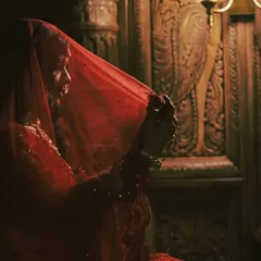 A classic bridal portrait using shadow and light. The bride, Swati, is captured in a moment of quiet reflection, her red veil creating a soft, ethereal glow around her in a grand, palace-like setting.