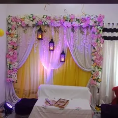 An intimate indoor Haldi decoration for a home setting. This arrangement uses layers of yellow and sheer white drapes, a floral border of pink and white flowers, and hanging lanterns to create a warm and cozy ambiance.