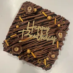 A close-up of my Ferrero Rocher brownie cake, perfect for a birthday celebration. It's drizzled with chocolate and topped with crunchy hazelnuts, sprinkles, and a "Happy Birthday" plaque.