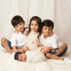 A beautiful sibling portrait where three older children in white and neutral tones are gathered lovingly around their new baby sibling, who is swaddled and sleeping peacefully on a soft white rug.