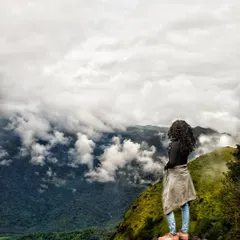 A traveler stands at the edge of a cliff, looking out over a valley filled with clouds and lush greenery. This photograph captures the immense scale and adventurous spirit of trekking in the Western Ghats.