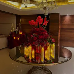 A grand floral centerpiece on a mirrored table, featuring a profusion of red flowers and surrounded by glowing electric candles. The reflection on the table and the chandelier above amplifies the sense of luxury.