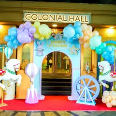 A whimsical pastel carnival entrance with carousel horses, hot air balloon props, and a beautiful balloon arch over the doorway.