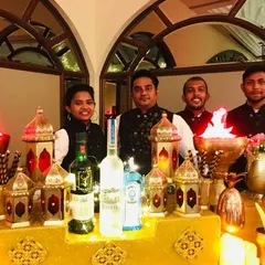 My team is ready to serve at this stunning Sufi night event. We stand behind a bar adorned with glowing lanterns and flame effects, dressed in elegant attire to complement the royal theme.