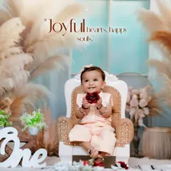 A happy baby girl holds a flower while sitting on a miniature throne. This studio session features a lovely boho theme with pampas grass and a "One" sign for her first birthday.