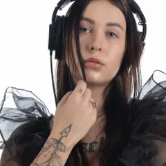A moody portrait of a female DJ with headphones, tattoos, and a sheer black top, captured against a clean white background.