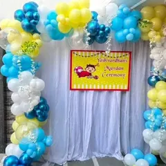 A colorful and festive balloon arch for a 'Mundan Ceremony'. The setup includes a custom banner with a cute graphic and a backdrop of white curtains.