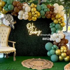 This beautiful 'Oh Baby' setup is perfect for a baby shower. It features a green hedge wall, a custom neon sign, and an organic balloon arch in earthy tones of green, brown, and gold.