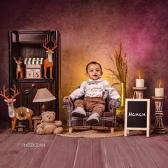 This rustic, woodland-themed studio setup is perfect for a classic toddler boy portrait. Dressed in a smart shirt and suspenders, he sits confidently in a scene filled with warm, earthy tones, deer figures, and soft candlelight.