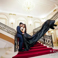 A couple stands on a grand staircase, her black trail gown flowing down the red carpet, creating a striking visual of elegance and drama for a pre-wedding shoot.
