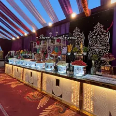 A magnificent dessert and fruit counter that serves as a visual masterpiece. The display is built on illuminated platforms with intricate silver and gold backdrops, featuring everything from fresh fruit to traditional Indian sweets.