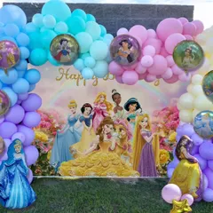 A dreamy Disney Princess themed birthday party setup for an outdoor celebration. The backdrop features all the popular princesses and is framed by a beautiful pastel rainbow balloon arch.