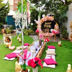 A bright and cheerful garden party with low picnic-style seating. The setup is decorated with pink floral arrangements, balloons, and personalized photos of the birthday girl.