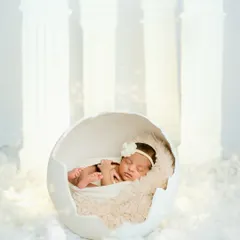 A heavenly themed newborn shoot. This little one is nestled inside a white eggshell prop, surrounded by soft clouds and ethereal white pillars, creating a truly angelic and dreamlike portrait.