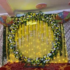 A vibrant and modern Haldi backdrop. This setup features a full circular arch covered in yellow and white flowers, with a custom 'Haldi' neon sign glowing in the center against a bright yellow drape and fairy lights.