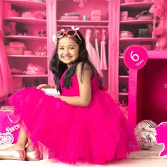 A six-year-old girl smiles brightly in her Barbie-themed shoot, sitting beside a life-sized Barbie box in a room filled with pink.