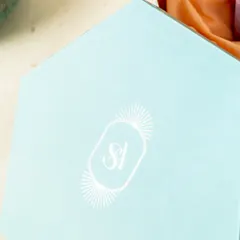 A close-up of the lid of the hexagonal invitation box, featuring the couple's elegant 'SA' monogram in a clean, white print against a powder blue background.