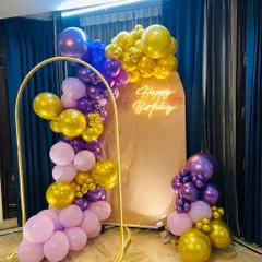 A modern and chic birthday setup at home. The decor includes a stylish balloon garland in purple, pink, and gold, arranged around a metal arch and a simple backdrop with a neon sign.