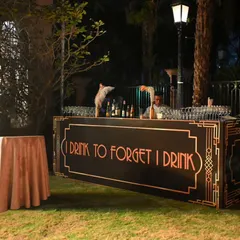 An outdoor bar setup for the Gatsby party. The custom-built bar front with its period-appropriate font and design, along with professional bartenders, added an authentic touch to the celebration.