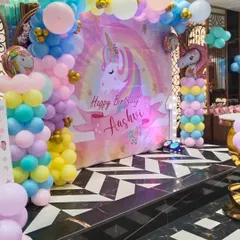 A magical first birthday party with a Unicorn theme. The decoration includes a pastel-colored balloon arch, a personalized backdrop with a unicorn graphic, and themed foil balloons.