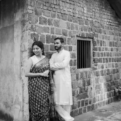 A timeless black and white portrait. The couple, dressed in traditional attire, stands by a rustic stone house, creating an image that feels like a cherished memory from another era.