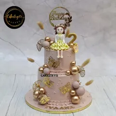 A sophisticated two-tier chocolate cake for a stylish second birthday. It's topped with a cute girl figurine and decorated with golden butterflies, delicate ferns, and shimmering faux pearls for an elegant touch.