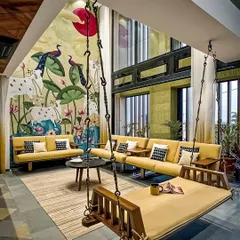 For clients who love art and tradition, we design unique living spaces. This room features a high ceiling with a stunning, large-scale Pichwai-style mural, complemented by simple yellow sofas and a traditional wooden swing.