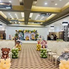 A full view of a banquet hall decorated for a jungle theme birthday. The setup includes a main stage backdrop, and the walkway is lined with balloon pillars topped with cute animal cutouts like bears, cats, and monkeys.