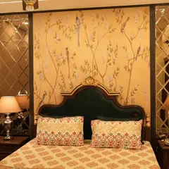 A bedroom with a vintage aesthetic, featuring a dark wood bed with a velvet headboard. The accent wall has a delicate, traditional wallpaper, and the look is completed with antique-style mirrored panels.