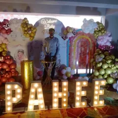 A grand 'Welcome Baby' setup featuring large marquee letters that spell out the baby's name, 'PAHEE'. The backdrop includes a neon sign, a rainbow arch, and balloon clouds for a spectacular welcome.