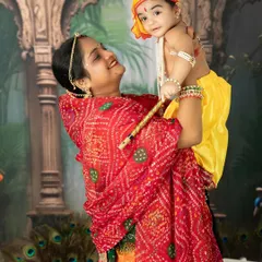 A mother dressed in a vibrant red bandhani saree joyfully lifts her baby, who is dressed as little Krishna, during a festive Janmashtami photoshoot.