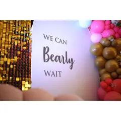 A close-up of the baby shower backdrop, clearly showing the theme "We Can Bearly Wait" next to a shimmering gold sequin panel.