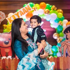 A mother holds her two-year-old son, both smiling, at his fun-filled birthday party.