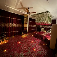 A full room view showing the impact of a ceiling installation. The floor is covered in red rose petals, leading to a bed under a canopy of hanging flowers and lights.