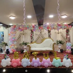 An elegant and grand first birthday setup in a party hall. This design includes a wide pastel-colored organic balloon arch over a regal white chaise lounge, with the name "BATUL" spelled out in balloon letters at the front.