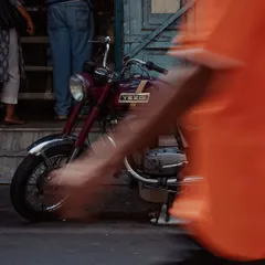 A vintage maroon Yezdi motorcycle is captured in sharp focus while the motion of a person walking past is blurred into an orange streak. This artistic technique freezes a moment in time, contrasting the timeless bike with the city's constant movement.