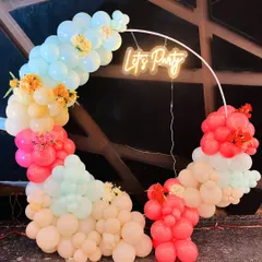 This circular balloon arch uses a soft, pastel color scheme and a "Let's Party" neon sign. It's a versatile setup that works well for daytime engagement parties, bridal showers, or any modern celebration.