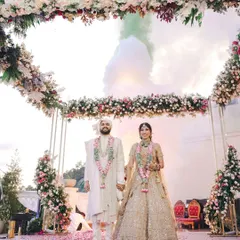 The couple stands hand-in-hand at their outdoor wedding ceremony, framed by a magnificent floral arch with a backdrop of misty mountains and colored smoke.