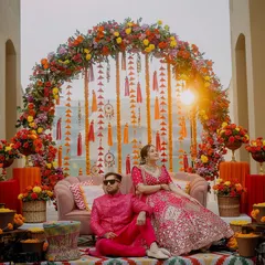 A regal portrait from a Mehendi ceremony. The couple is seated against a stunning floral arch, the golden hour sunlight adding a warm glow to the vibrant, colorful decor.
