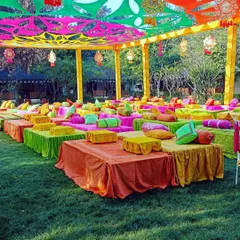 A vibrant outdoor Mehendi setup with low seating and colorful cushions under a canopy of bright orange, pink, and green drapes.