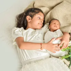A tender moment between siblings, as an older sister lies down and gently holds her swaddled newborn sibling, looking at them with love and wonder.