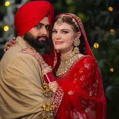 This portrait from a beautiful NRI wedding showcases the couple's regal attire and their comfortable connection. The bride's traditional red lehenga and the groom's turban create a stunning visual against the soft, natural background.