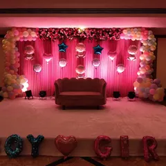 An elegant stage setup for a baby shower, using soft pink lighting and drapes to create a warm ambiance. The decoration includes a pastel balloon arch, a floral border, and hanging foil balloons, with large 'BOY' or 'GIRL' foil letters in front, making it perfect for a gender reveal party.