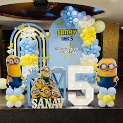 A fun-filled Minions theme for a 5th birthday party. The setup includes multiple Minion character cutouts, a large light-up number '5', and a blue and yellow balloon arch decorated with bananas.