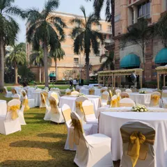 Guest seating for a sunny outdoor luncheon. The round tables are covered in white linen, and the chairs are decorated with golden sashes, creating a cheerful and elegant look.